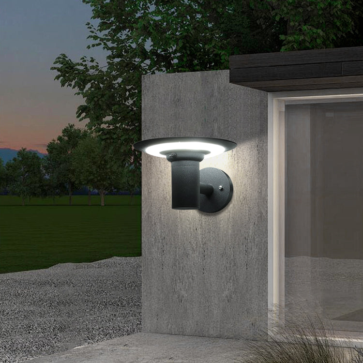 Outdoor wall light fixture on a concrete pillar with a garden and trees in the background.